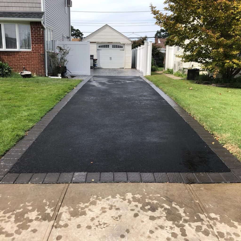 A freshly paved asphalt driveway, crafted by a skilled masonry contractor in Nassau County, leads to a white garage. It is bordered by bricks, flanked by green grass and a few plants. A house is partially visible on the left, while a white fence lines the right side under an overcast sky.