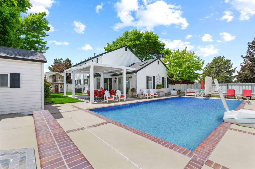 The backyard boasts a rectangular swimming pool framed by a patio with brick accents. A white house sits in the background, complemented by outdoor seating with red cushions under a covered patio. Lush greenery and trees add to the charm, showcasing the fine work of a masonry contractor from Suffolk County on this sunny day.