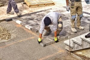 A construction worker wearing gloves and knee pads is placing rectangular pavers on a gravel base, building a pathway for an outdoor kitchen in Suffolk County. Another worker stands nearby, while various tools and materials are visible in the background.