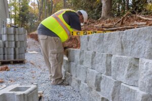 A construction worker in a neon vest and cap, representing a skilled masonry contractor from Suffolk County, is diligently building a stone retaining wall outdoors. Using a level for precise alignment, the wall rises on a gravel surface surrounded by trees and a partially cleared area.