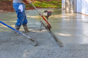 A construction worker smooths wet concrete with a power trowel on a driveway, possibly laid by a paving company in Nassau County. The person is wearing jeans, a gray shirt, and boots. A second tool is visible, and sunlight highlights the work area in front of a garage.