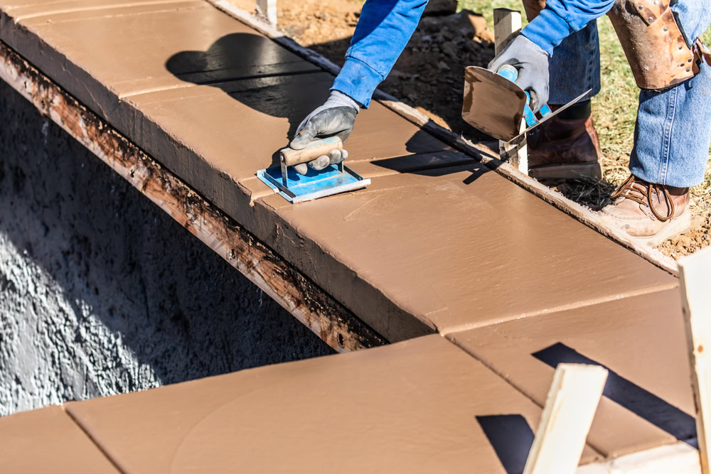 A construction worker in blue clothes and gloves smooths wet concrete on a wall corner with a trowel, crafting the foundation for an outdoor kitchen in Suffolk County. Sunlight casts shadows as tools and materials are scattered around the work area, hinting at the project's scale.