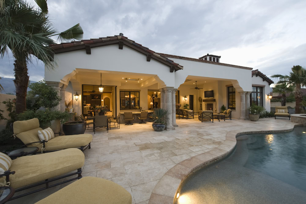 A luxurious backyard features a Mediterranean-style house with an open terrace, lit by warm lights. An outdoor kitchen crafted by a masonry contractor in Nassau County complements the cushioned lounge chairs, landscaped garden, and smoothly curved pool under a cloudy sky.