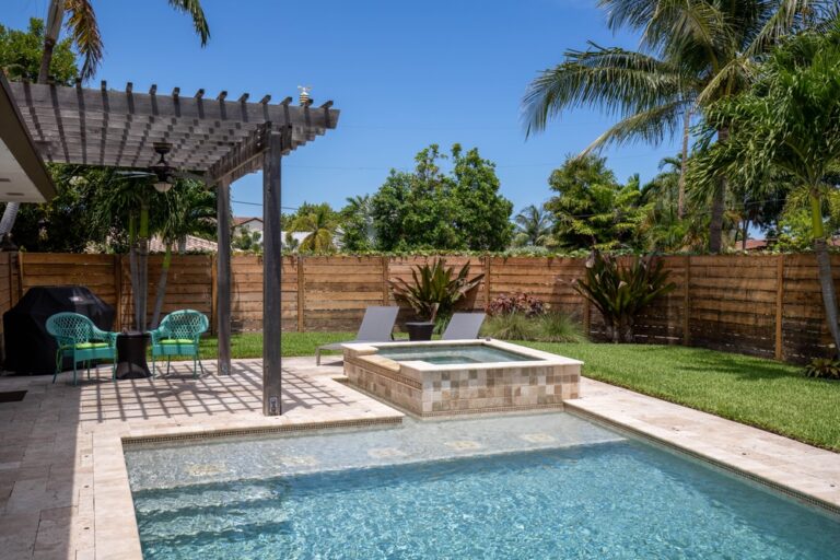 A backyard oasis with a swimming pool and hot tub features two lounge chairs and a small table with green chairs under a pergola. Tropical plants and a wooden fence provide privacy, while an outdoor kitchen installed by an expert masonry contractor in Suffolk County completes the serene setting.