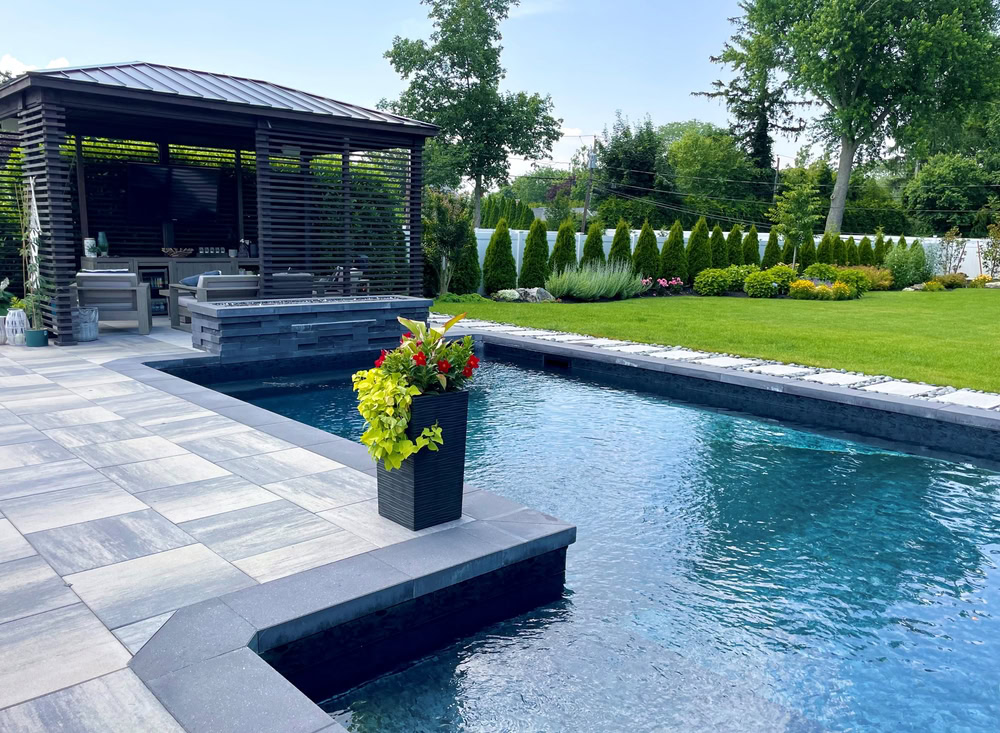 A modern backyard features a rectangular pool with blue water, surrounded by gray stone tiles courtesy of a paving company in Nassau County. A pergola with seating is in the background, and lush green grass and shrubs border the area. A planter with yellow and red flowers decorates the poolside.