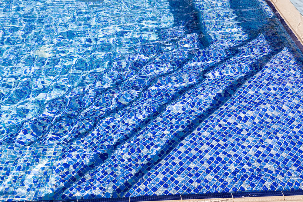 Blue swimming pool with mosaic tile steps leading into the water, reflecting light and creating a rippling effect on the surface, perfectly complementing your outdoor kitchen in Nassau County.