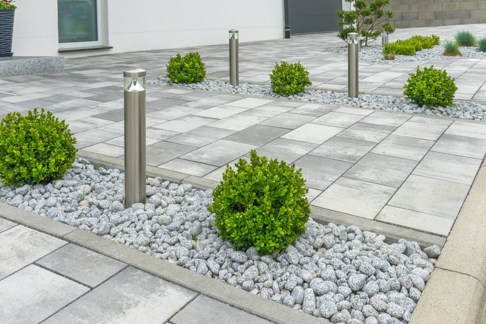 Modern outdoor walkway with gray concrete tiles, bordered by round green shrubs and small gray stones. Metal bollard lights are placed strategically along the path, enhancing the minimalist landscape design in front of a white building. Perfect for an Outdoor Kitchen Suffolk County installation.