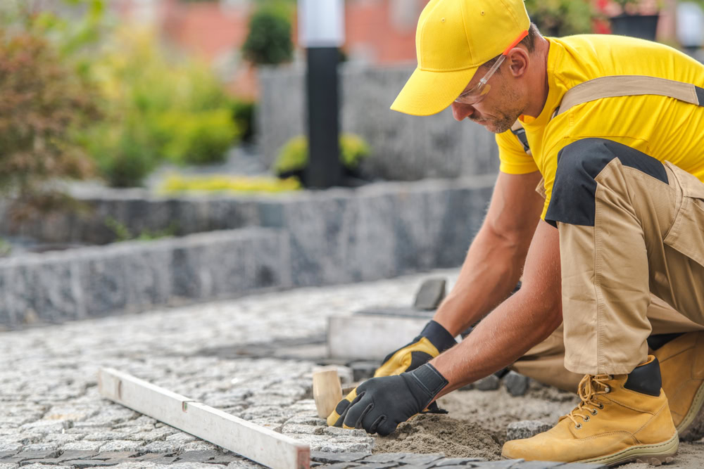 A person from a renowned Paving Company in Nassau County is wearing a yellow cap and shirt with beige overalls, kneeling to lay gray cobblestones outdoors. They’re equipped with gloves and protective glasses, while blurred plants and steps add depth to the background.