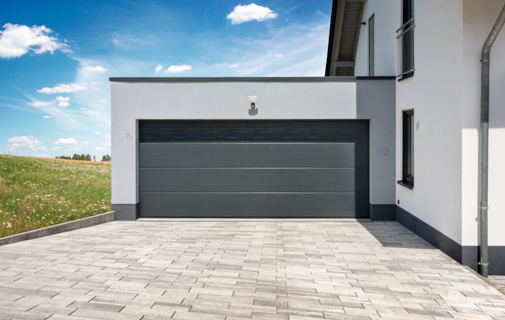 A modern house boasts a sleek grey garage door and a neatly paved driveway crafted by a renowned Paving Company in Suffolk County. The building's exterior is white, under a clear sky with scattered clouds, and to the left lies a verdant grassy field.