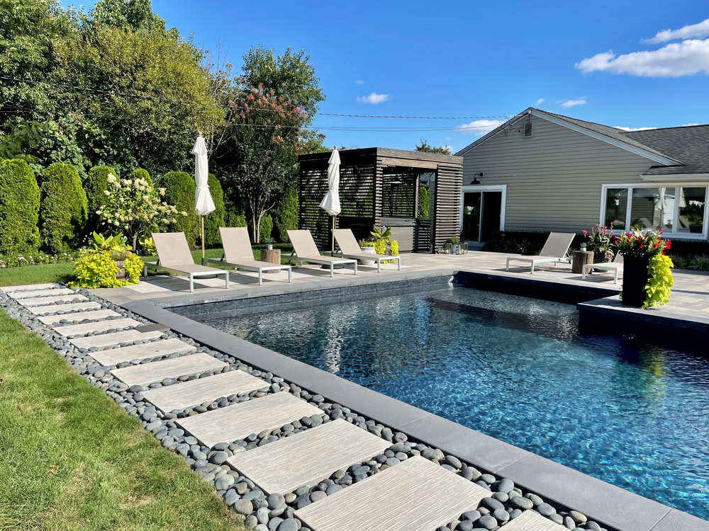 A backyard with a modern rectangular pool surrounded by a stone path and loungers under umbrellas. A wooden pergola is in the background near a single-story house. The sky is clear and blue, and greenery surrounds the area.