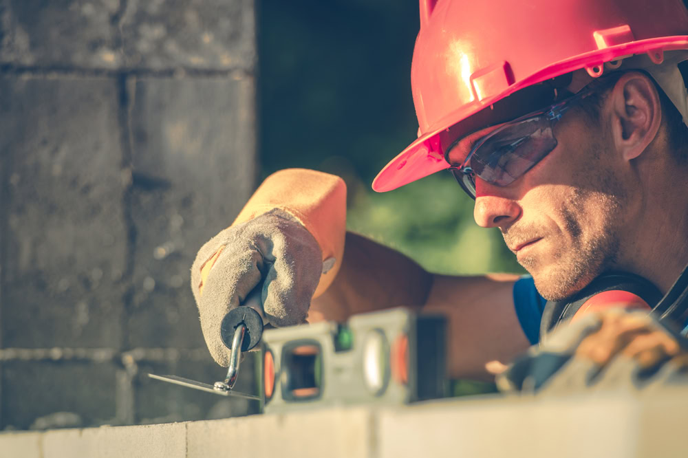 A construction worker wearing a red hard hat and safety glasses is using a spirit level on a concrete block. The worker is focused, holding the level with gloved hands. The background is blurred, highlighting the worker and tools.