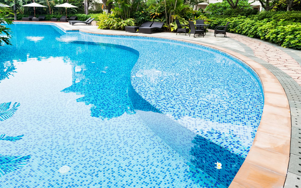 A serene swimming pool with clear blue water surrounded by lush greenery and poolside loungers. The pool features a mosaic tile design, and the area is tranquil, with some umbrellas providing shade in the background.