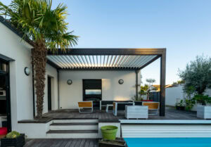 A modern wooden deck with a pergola featuring adjustable slats. The deck has lounge chairs, potted plants, and overlooks a swimming pool. A tall palm tree is on the left, and the house has white walls and black-framed windows.