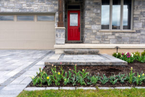 Front of a stone house with a red door, three steps leading to the entrance, and a gray driveway. Tulip garden with colorful flowers lines the walkway.