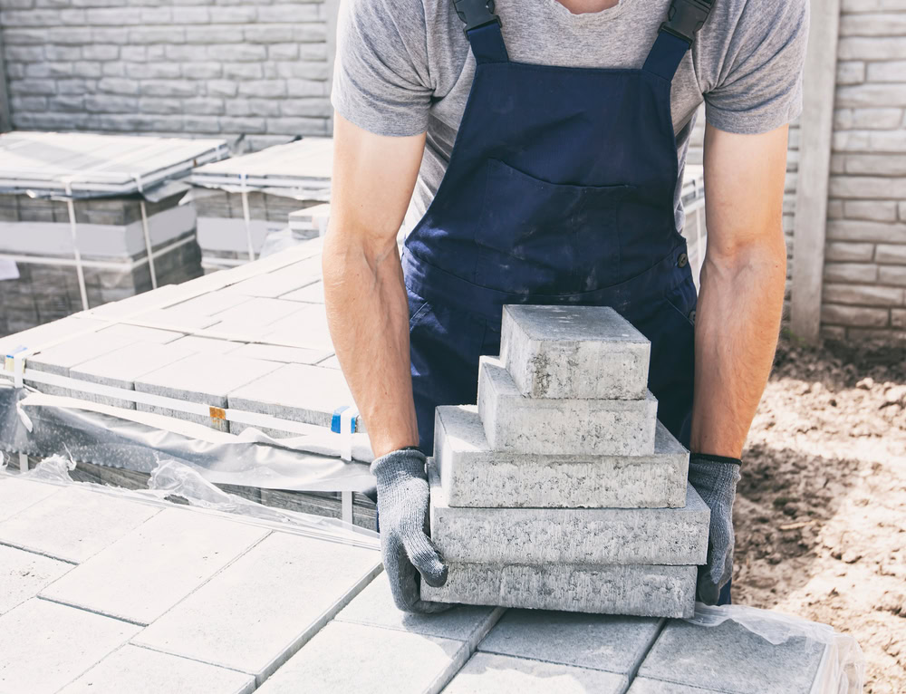 A worker in navy overalls and gloves is arranging gray concrete slabs in a tiered formation at a construction site. The background shows neatly stacked concrete blocks and a stone wall.