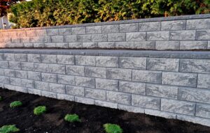 A two-tiered gray stone retaining wall with horizontal layers is shown. Small green shrubs are planted in the soil at the base, and dense green foliage tops the wall. The scene is evenly lit, suggesting late afternoon sunlight.