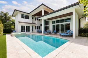 A modern two-story white house features large windows and a spacious backyard with a rectangular swimming pool surrounded by stone tiles. Blue cushioned patio furniture decorates the poolside under a covered patio, and greenery surrounds the area.