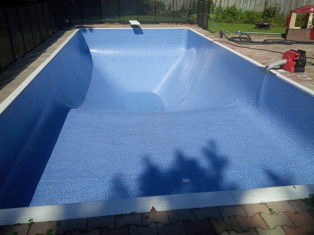 Empty swimming pool with blue liner, surrounded by a brick patio and black fence. A red vacuum-like machine is on the right edge, and there are shadowy trees in the foreground.