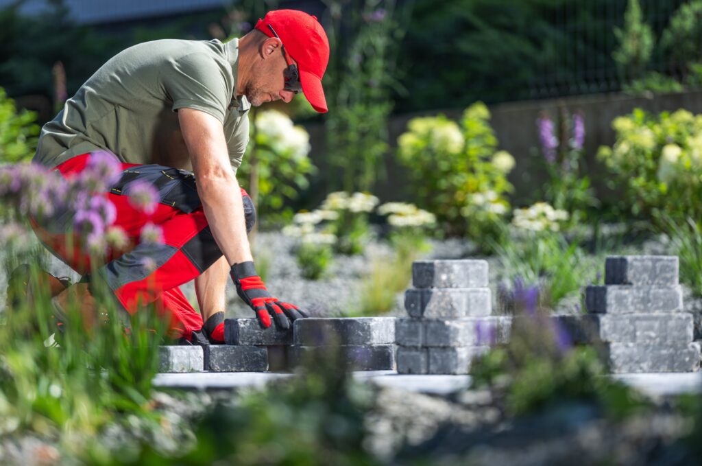 A worker wearing a red cap and gloves is kneeling while arranging gray bricks in a garden. The background features green foliage and blooming flowers, adding a vibrant touch to the scene.