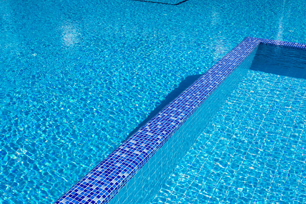 A clear blue swimming pool with a section of the wall visible, adorned in small, square blue tiles. Sunlight reflects on the water, creating shimmering patterns on the surface.