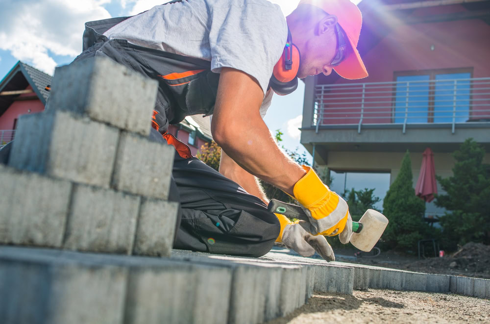 A worker wearing a red cap, orange ear protection, and gloves is kneeling and placing concrete pavers with a rubber mallet. The scene is outdoors near a modern house with a blue sky and clouds in the background.