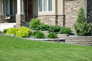 A well-maintained front yard featuring a lush green lawn, tiered stone landscaping with vibrant green shrubs, and a stone facade house with a porch.